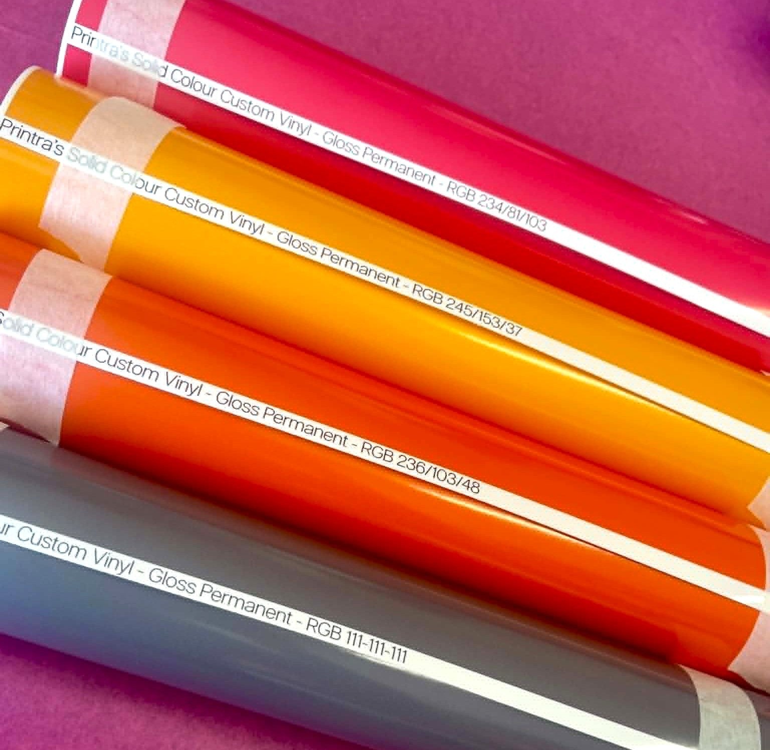 Four rolls of glossy custom printed vinyl in red, orange, yellow, and grey, arranged diagonally on a purple background. White paper labels with black text are visible on each roll.