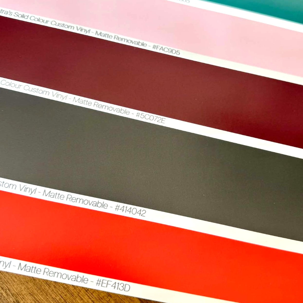A close-up of custom printed vinyl colour swatches with text labels including 'Matte Removable' and colour codes, showing diagonal bands of pink, burgundy, dark grey, and bright red orange.