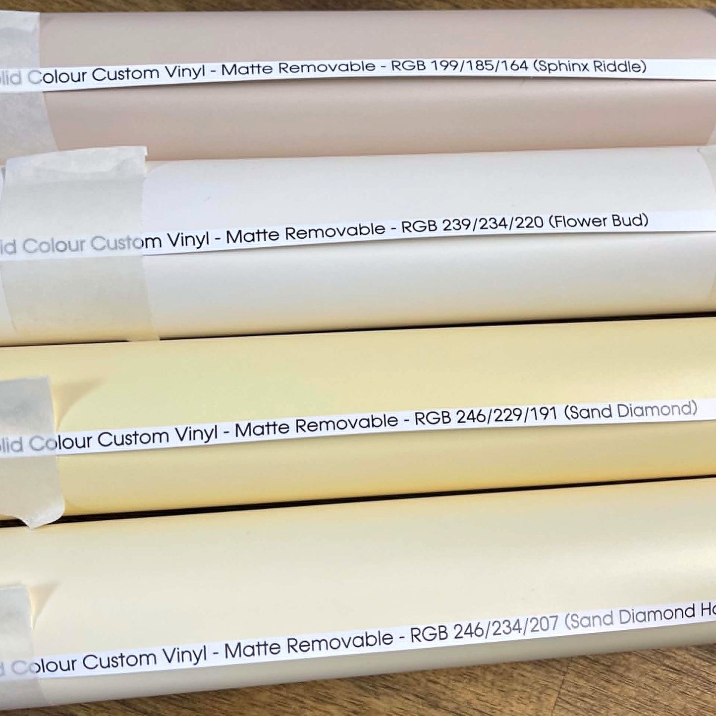 Several rolls of custom printed matte removable vinyl stacked horizontally, each labelled with a different colour name and RGB values, including Pink, Flower Bud, Sand Diamond, and others.