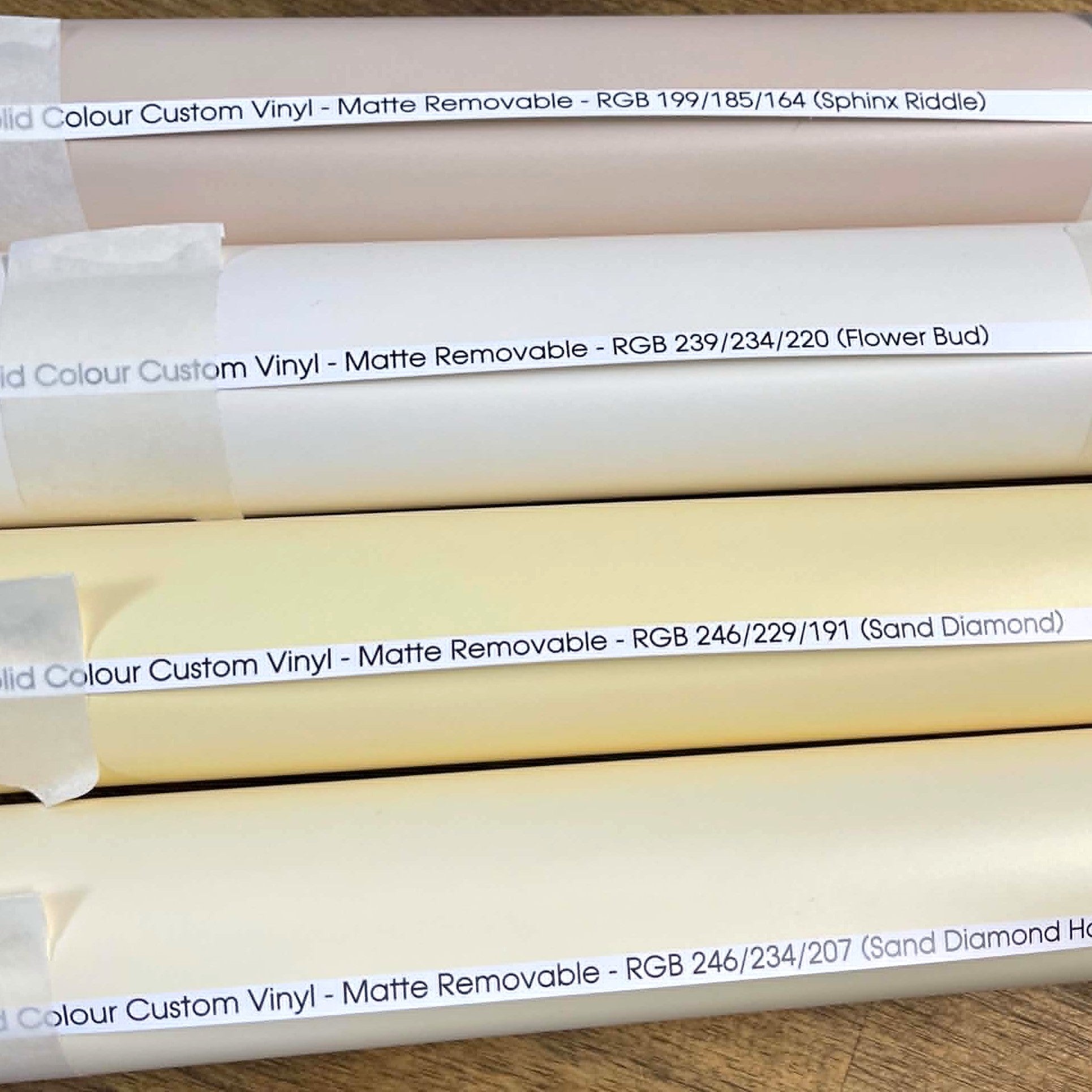 Several rolls of custom printed matte removable vinyl stacked horizontally, each labelled with a different colour name and RGB values, including Pink, Flower Bud, Sand Diamond, and others.